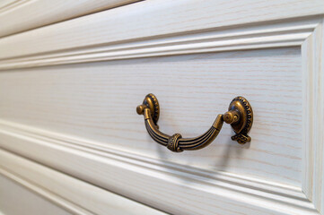 A bronze color handle on a white dresser. The handle is ornate