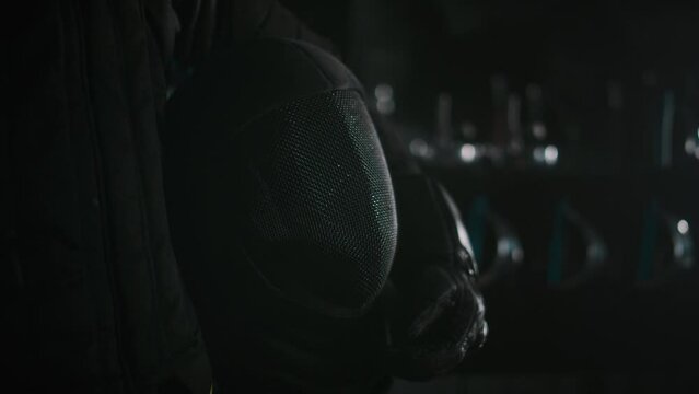 Midsection closeup of anonymous fencer in black uniform holding black epee mask in dark room