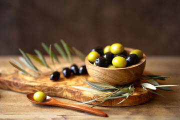 A bowl full of green and black olives on a rustic wooden table and a spoon with an olive. Nearby is an olive tree branch with olives and green leaves