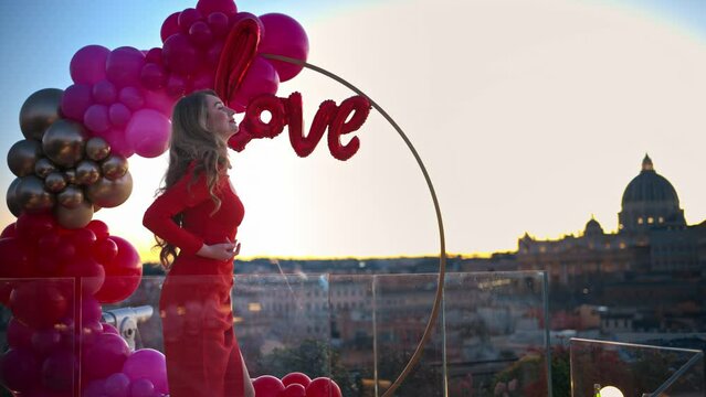 Beautiful young woman in red dress dancing at sunset near the red and pink balloon arch spelling out the word "love". Vatican city in background. Rome, Italy