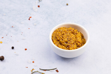 grainy mustard in a white ceramic bowl on a blue table surrounded by spices, herbs and salt