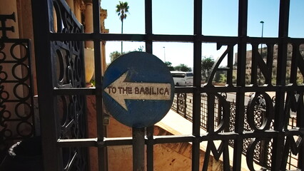 Directional Sign To The Basilica Through Wrought Iron Gate, Sunny Day Perspective