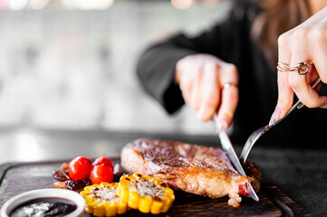 close-up of someone slicing a grilled steak. The well-cooked steak, accompanied by grilled corn and roasted tomatoes, is presented on a dark wooden cutting board.