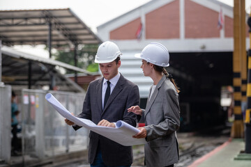 Supervisors architect and engineers wearing safety white helmet ้reading blueprint railway track design. Inspecting station at train maintenance repair department factory.