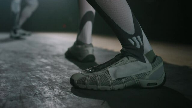 Ground level tracking footage of legs of unrecognizable fencer making few steps and taking crouching position before duel with opponent in dark hall