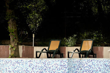 Side view of black and brown plastic sun loungers for relaxing on the hotel territory, backyard surrounded by trees, park, mosaic tile. Background with copy space. Concept of vacation, sunbathing.