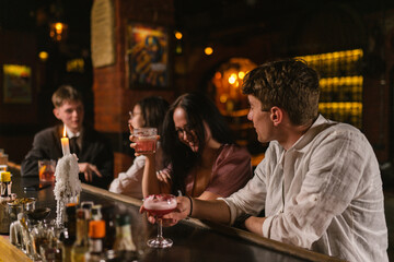 Guy in classic shirt tells jokes to brunette girl in glasses with alcoholic drink. Young people at...