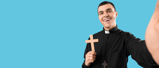 Young priest with cross taking selfie on blue background, closeup