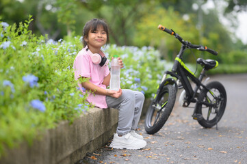A young girl sits and drinks water next to a bicycle in the park.