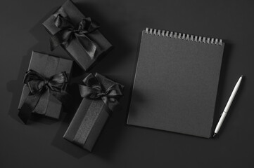 Black gift boxes with satin ribbons on a black background and a black notepad with a white pen....