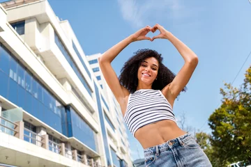 Rolgordijnen Photo of cheerful lovely pretty girl wear crop top stands with raised hands demonstrate heart sign sun blue sky outdoors © deagreez