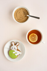 Gingerbread in the shape of a rabbit with an Easter egg on a plate, a cup of tea with a slice of lemon, a sugar bowl with cane sugar on a light table. Top view.
