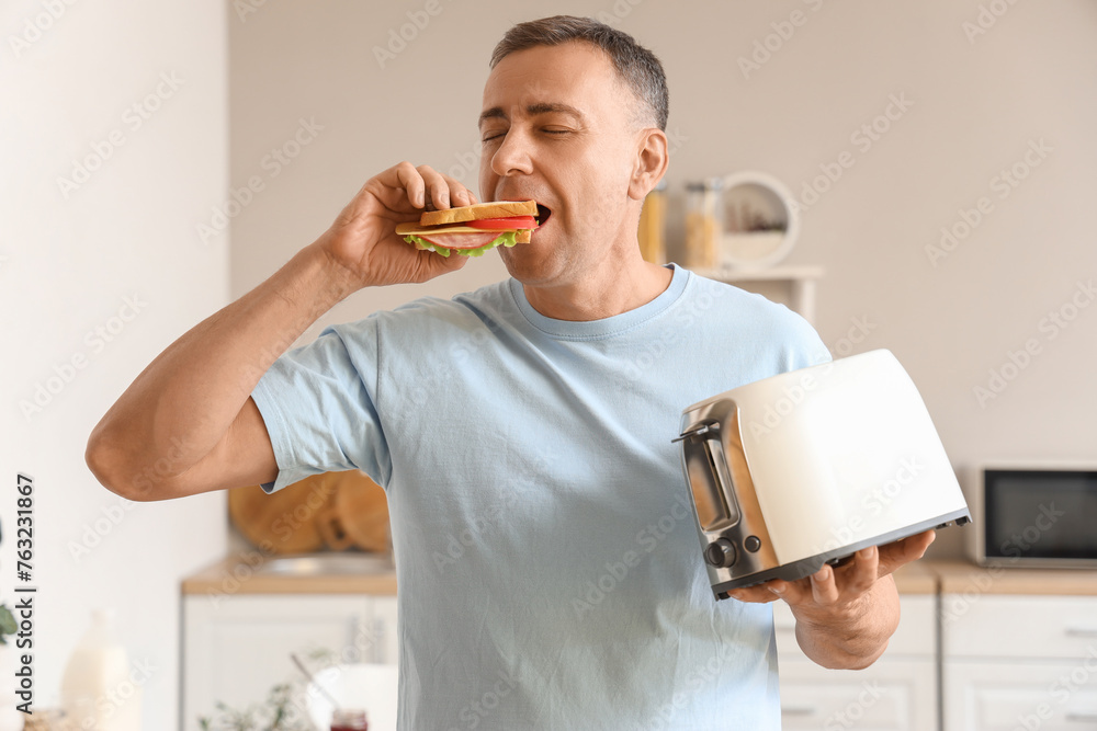 Canvas Prints Middle-aged man with toaster eating delicious sandwich in kitchen