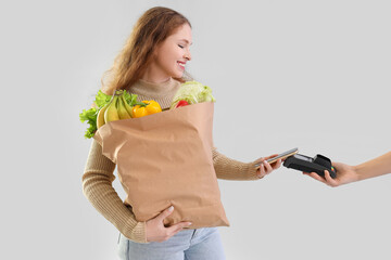 Young woman with paper bag of healthy food paying via terminal on light background