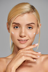 portrait alluring young woman with cream on cheeks touching her chin against gray background