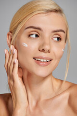 beauty girl in her 20s applying cream on her cheeks and looking to side against grey background