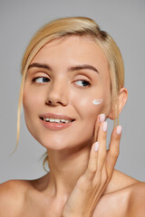 portrait pretty girl in her 20s with healthy skin applying cream on face in grey background