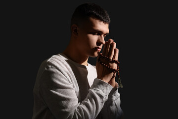 Young man with rosary beads praying on dark background