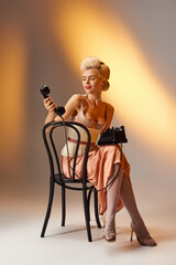 alluring blonde young woman in corset dress posing on chair with retro phone against gray background