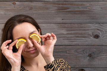 Portrait of a young white woman standing in front of a wall made of old wooden boards and holding lemons half-slices over her eyes. 