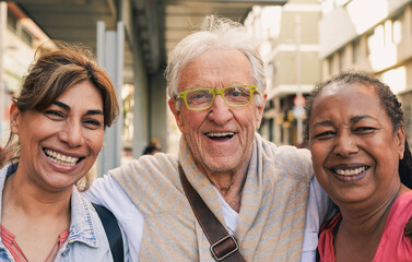 Multiracial senior people hugging each other while smiling on camera with city in background - Joyful elderly friendship and pension concept
