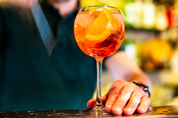 vibrant orange cocktail served by a bartender. The drink, garnished with a slice of orange, stands out against the darker background. bartender’s hand is visible, and the setting appears to be a bar.