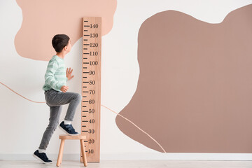 Cute little boy on stool measuring height near wooden stadiometer at home