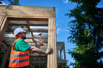 Carpenter constructing wooden frame house near the forest. Bearded man hammering nails into...