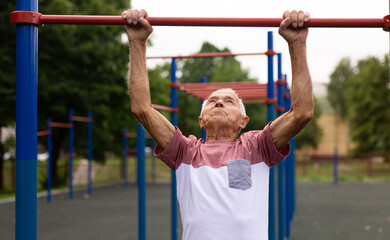 Old man pulls himself up on horizontal bar on sports ground. Outdoor sports in old age