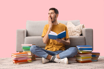 Sleepy young man with books sitting near sofa on pink background