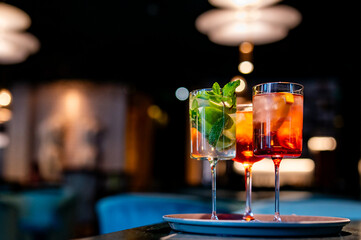 Two vibrant cocktails adorned with mint leaves rest on a tray in an elegant, softly lit bar setting. One is a green mojito, the other a reddish-orange concoction, both served in long-stemmed glasses