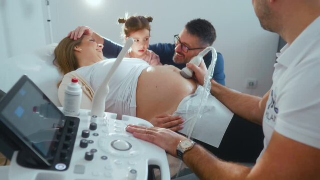 Husband and little daughter provide support to pregnant mother who is lying on medical couch during ultrasound scanning