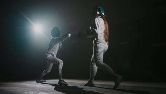 Back full footage of two foilswomen in protective gear attacking each other with rapiers during duel in dark hall