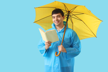 Young man in raincoat with umbrella reading book on blue background