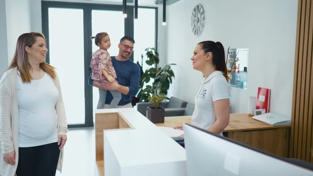 A pregnant woman with her husband and little daughter visits a medical clinic for a gynecological examination. Friendly female hospital receptionist talking to clients at the reception desk in a medic