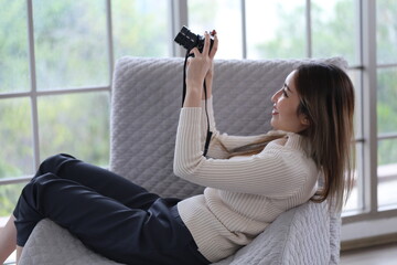 Asian woman lying on sofa holding DSLR camera, reviewing photos from camera before sending photos...