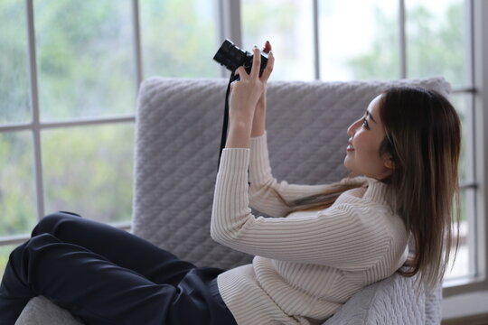Asian woman lying on sofa holding DSLR camera, reviewing photos from camera before sending photos for sale in apartment