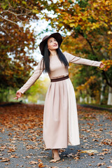 Cute young woman with wide-brimmed hat and fashionable attire standing on pathway surrounded by fall trees