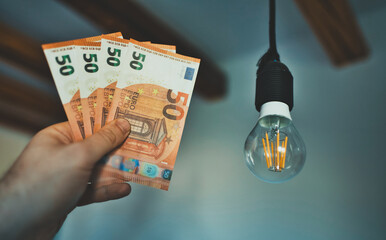 Man holds money next to a light bulb. Expensive electricity concept. - 763285417