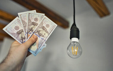Man holds money next to a light bulb. Expensive electricity concept. - 763285418