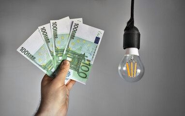 Man holds money next to a light bulb. Expensive electricity concept. - 763285423