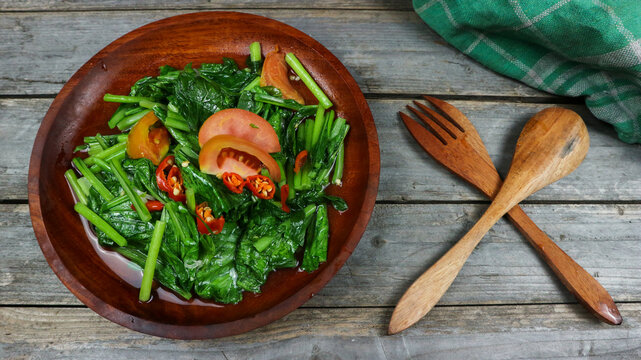 Home made food green mustard vegetables or tumis sayur sawi with sliced chilli and tomatoes on a wooden table. Grey background.