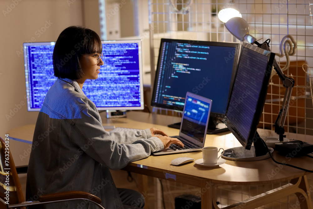 Poster female programmer with laptop and computer monitors working in office at night