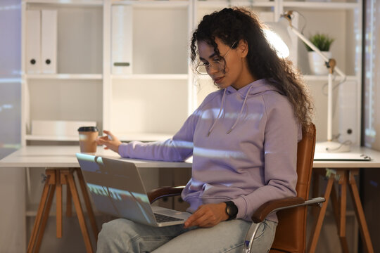 Female African-American programmer with laptop taking coffee cup from table in office at night