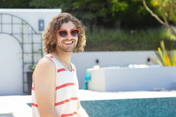 A young Caucasian man smiles by pool, wearing sunglasses, with copy space at home
