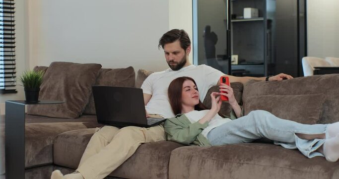 Happy family couple relaxing on sofa, man working on laptop, woman, wife, kneeling on man, scrolling social media on mobile phone at home. Concept of remote work.