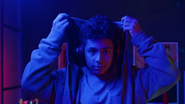 Chest up slowmo portrait of Biracial teenage gamer boy standing in neon blue room looking at camera while putting on hood