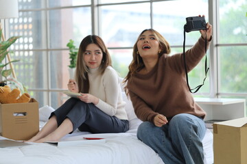 Two Asian female roommates sit on a bed together, one holds a camera and takes a selfie together, while fellow writers document a good relationship in an apartment room