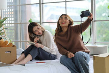 Two Asian female roommates sit on a bed together, one holds a camera and takes a selfie together,...