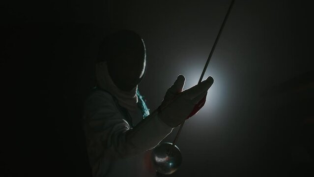 Medium footage of foilsman in epee mask holding rapier and looking at its blade while standing against lightMedium footage of foilsman in epee mask holding rapier and looking at its blade while standi